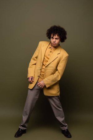 longitud completa del hombre afroamericano de moda en pantalones y chaqueta amarilla posando sobre fondo verde gris