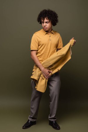 longitud completa de hombre afroamericano joven y de moda en polo amarillo sosteniendo chaqueta sobre fondo gris oliva