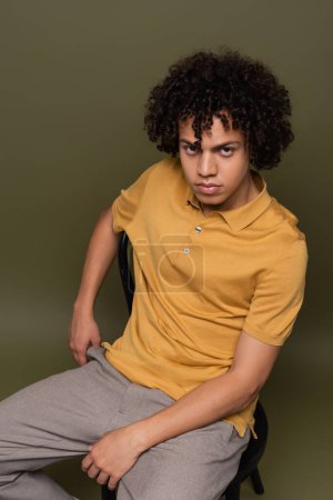 joven afroamericano hombre de polo amarillo sentado en silla y mirando a la cámara sobre fondo verde gris