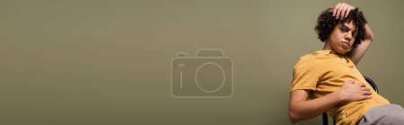 Foto de Hombre afroamericano de moda en polo amarillo agarrado de la mano en la cabeza y mirando a la cámara mientras está sentado aislado en gris, pancarta - Imagen libre de derechos