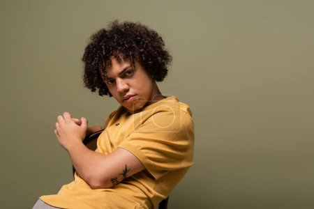 joven hombre afroamericano tatuado en polo amarillo mirando a la cámara mientras posa en silla aislada en gris