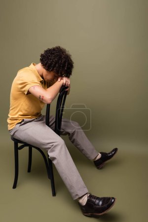 full length of tattooed african american man in yellow polo shirt and pants sitting on chair with bowed head on grey green background