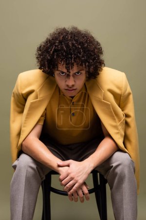 joven y confiado hombre afroamericano en chaqueta amarilla mirando a la cámara mientras está sentado aislado en gris
