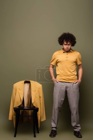 longitud completa del joven afroamericano de pie con las manos en los bolsillos cerca de la silla con chaqueta amarilla sobre fondo verde gris