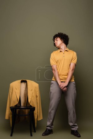 longitud completa del hombre afroamericano en polo amarillo y pantalones de pie con los ojos cerrados cerca de la silla con chaqueta sobre fondo verde gris
