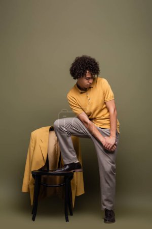 hombre afroamericano de moda pisando silla con chaqueta amarilla mientras posa sobre fondo verde gris