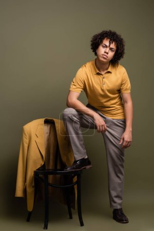 joven y elegante hombre afroamericano en camisa de polo y pantalones pisando silla cerca de chaqueta amarilla sobre fondo verde gris