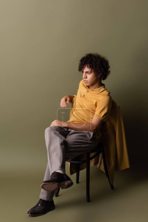 longitud completa de hombre afroamericano en polo amarillo sentado con las piernas cruzadas y mirando hacia otro lado sobre fondo verde gris