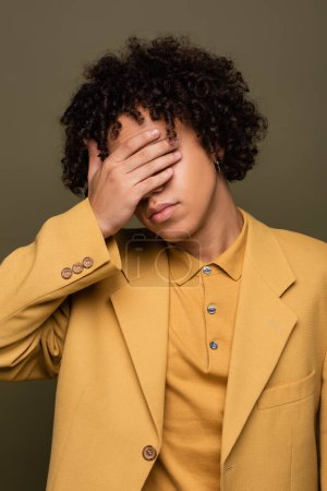 Téléchargez les photos : Homme afro-américain à la mode dans des vêtements élégants jaune couvrant visage avec la main isolé sur gris - en image libre de droit
