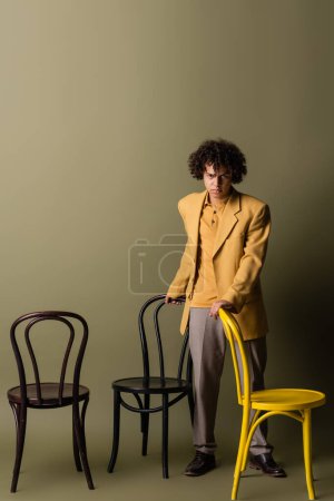 longitud completa de chico afroamericano en traje casual de moda de pie cerca de sillas negras y amarillas sobre fondo gris oliva