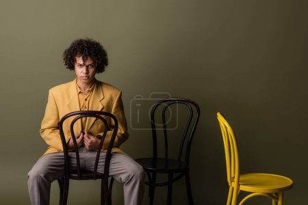 chico afroamericano en traje elegante posando con sillas negras y amarillas sobre fondo gris oliva