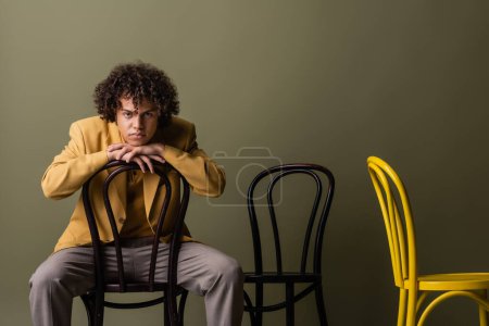 Photo for Brunette african american man in trendy casual outfit sitting on black chair and looking at camera on olive grey background - Royalty Free Image
