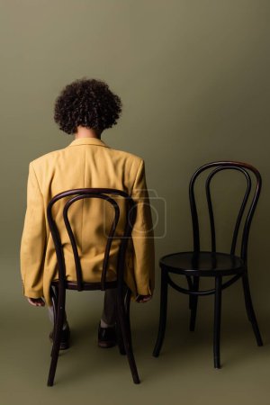 back view of brunette african american man with curly hair sitting in yellow blazer on black chair on grey green background