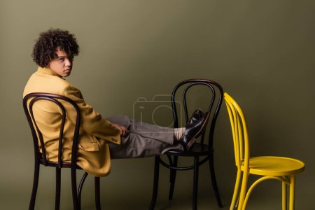 full length of stylish african american man in yellow blazer sitting on black chairs on grey green background