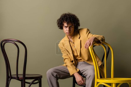 hombre afroamericano de moda en blazer y pantalones sentados cerca de la silla amarilla sobre fondo verde gris