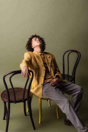 joven y moderno hombre afroamericano con los ojos cerrados sentado cerca de sillas negras sobre fondo gris oliva