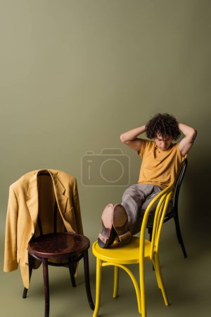 full length of stylish african american guy with hands behind head sitting on black and yellow chairs on grey green background