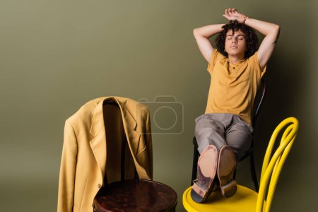 junger afrikanisch-amerikanischer Mann mit geschlossenen Augen und Händen hinter dem Kopf, der auf Stühlen in der Nähe des trendigen gelben Blazers auf graugrünem Hintergrund sitzt
