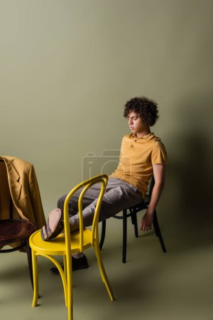 longitud completa del hombre afroamericano con los ojos cerrados sentado en sillas negras y amarillas cerca de chaqueta de moda sobre fondo verde gris