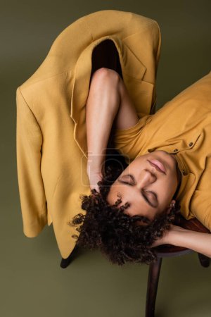 Foto de Vista de ángulo alto del hombre afroamericano acostado con los ojos cerrados y las manos detrás de la cabeza cerca de chaqueta amarilla en la silla sobre fondo verde gris - Imagen libre de derechos