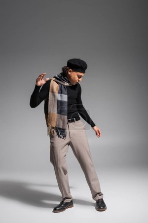 full length of stylish african american man in black sweater and plaid scarf with beret posing on grey background