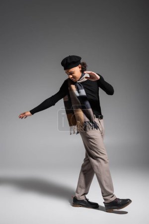 Foto de Full length of young and stylish african american man in plaid scarf and beret stepping on grey background - Imagen libre de derechos