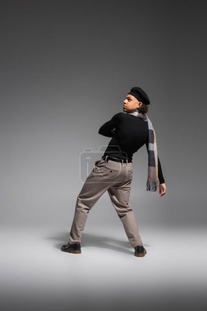 Foto de Longitud completa de hombre afroamericano en boina negra y cuello alto con bufanda a cuadros mirando hacia otro lado sobre fondo gris - Imagen libre de derechos