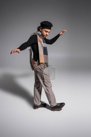 full length of african american man in black pullover with beret and plaid scarf stepping on grey background