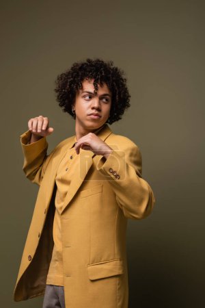 hombre afroamericano de moda en chaqueta amarilla de moda mirando hacia otro lado mientras está de pie sobre fondo gris