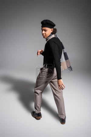 Foto de Longitud completa de hombre afroamericano joven y de moda en boina negra y jersey con bufanda cálida mirando a la cámara sobre fondo gris - Imagen libre de derechos
