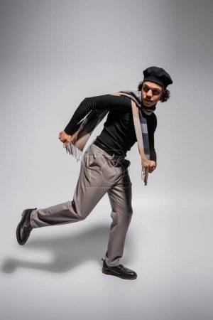 full length of african american man in black beret and sweater with trousers running and looking away on grey background