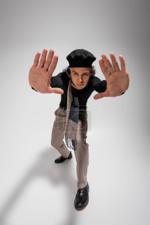 high angle view of african american man in beret and stylish casual attire gesturing with raised hands on grey background