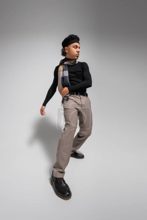 Foto de Chico afroamericano de moda en boina negra y suéter con bufanda mirando hacia otro lado sobre fondo gris - Imagen libre de derechos