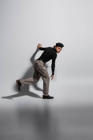 Téléchargez les photos : Full length of young african american man in black turtleneck and trendy beret looking at camera while walking on grey background - en image libre de droit