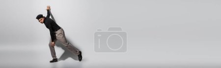 full length of trendy african american man in black pullover and pants posing on grey background with shadow, banner