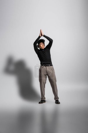 Afrikanisch-amerikanischer Mann in schwarzer Baskenmütze und Pullover posiert mit erhobenen betenden Händen auf grauem Hintergrund