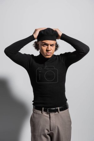 Foto de Frowning african american man in black beret and turtleneck with trousers standing with hands on head on grey background - Imagen libre de derechos