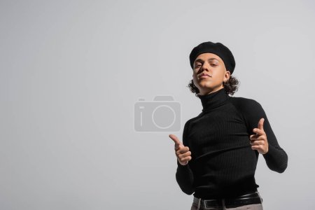 trendiger afrikanisch-amerikanischer Mann in schwarzem Pullover und Baskenmütze zeigt Fingergewehr-Geste, während er isoliert auf grau in die Kamera blickt