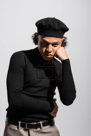 young and trendy african american man in black beret and turtleneck standing with hand near face and looking at camera isolated on grey