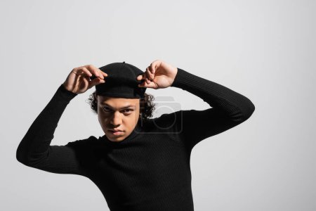 stylish african american man in black turtleneck touching beret and looking at camera isolated on grey