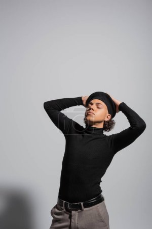 african american guy in black pullover and beret posing with closed eyes and hands on head on grey background