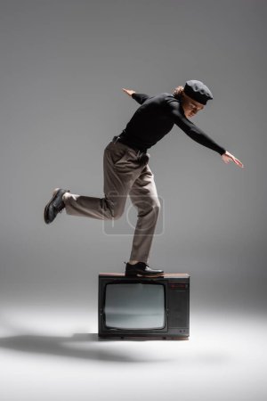 full length of trendy african american man in black sweater and trousers balancing on vintage tv set on grey background