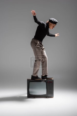 full length of african american man in trendy casual attire balancing on vintage tv set on grey background