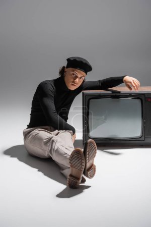 full length of african american man in trendy beret and black turtleneck sitting near vintage tv set and looking at camera on grey background