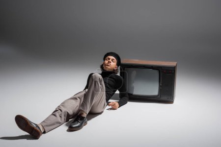 voller Länge junger afrikanisch-amerikanischer Mann mit geschlossenen Augen, der in Pullover und Hose in der Nähe von Retro-TV auf grauem Hintergrund liegt