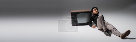 Foto de Full length of african american man in black beret and trousers leaning on vintage tv set while lying on grey background, banner - Imagen libre de derechos