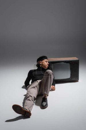 full length of fashionable african american man in black turtleneck and beret lying near vintage tv set and looking away on grey 