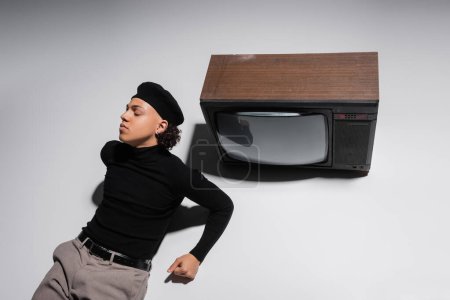 high angle view of african american man in black beret and turtleneck posing near vintage tv set on grey background