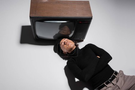 top view of african american man in black sweater and beret lying with closed eyes and crossed arms near vintage tv set on grey 