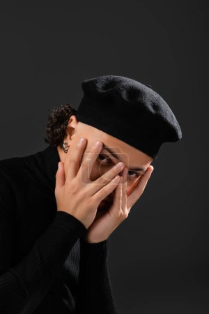 Foto de Joven afroamericano hombre en boina de moda oscureciendo la cara con las manos y mirando a la cámara aislada en negro - Imagen libre de derechos